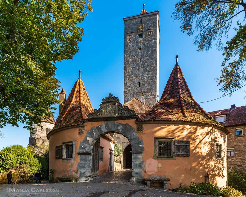Rothenburg ob der Tauber - Zusehen ist das dickbäuchige Burgtor in organge mit dem hohen kantigen Burgturm und Stadttor aus alten grauen Steinen. Das Sonnenlicht scheint quer von rechts auf den Turm. Vom vorderen Turm sieht man den Durchgang und die Brücke zum hinteren Turm. 