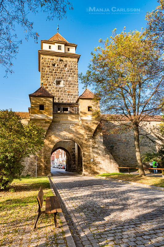 Rothenburg ob der Tauber - Galgentor mit Parkbank links davor und eine Straße aus Kopfsteinpflanster, die durch das Tor führt. Der Turm besteht aus einem hohen eckigen Turm und an den Seiten sind kleine Turmspitzen angesetzt.