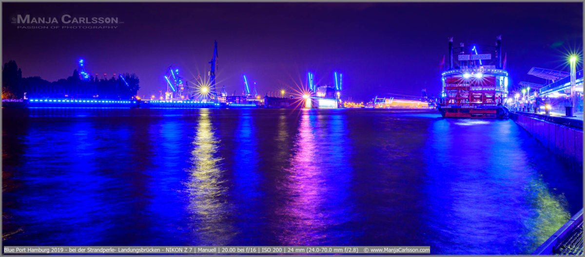 Blue Port Hamburg 2019 - Landungsbrücken mit Blick auf die Trockendocks von Blohm + Voss und dem großen Hafenrundfahrtschiff mit großen Schaufelrad am Heck. Der Himmel und das Elbwasser sind durch den Blue Port blau. Die Hafenbeleuchtung wirft farbige Lichtrefelktionen aufs Wasser.